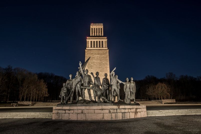 Die Figurengruppe am Mahnmal Buchenwald vor dem Glockenturm der Mahnmalsanlage bei Nacht. Sowohl Figurengruppe als auch Glockenturm sind angestrahlt.